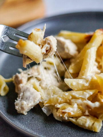 Fork holding up bite of ziti pasta, baked chicken, alfredo sauce and stringy melted cheese.