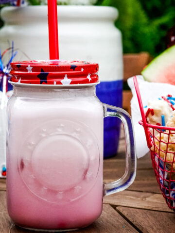 Frosted lemonade in a glass jar with a handle and a red white and blue lid with a straw.