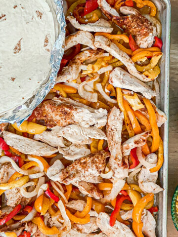 Sheetpan of cooked fajitas with slices of seasoned chicken, sliced onions and red and orange bell peppers with a stack of flour tortillas on top. Bowls of shredded cheddar cheese, sour cream and avocado on the side.