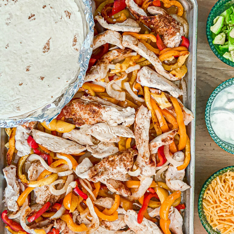 Sheetpan of cooked fajitas with slices of seasoned chicken, sliced onions and red and orange bell peppers with a stack of flour tortillas on top. Bowls of shredded cheddar cheese, sour cream and avocado on the side.