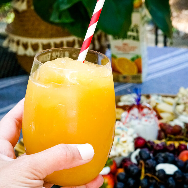 Hand holding orange juice bourbon slushie in clear glass with red and white paper straw.