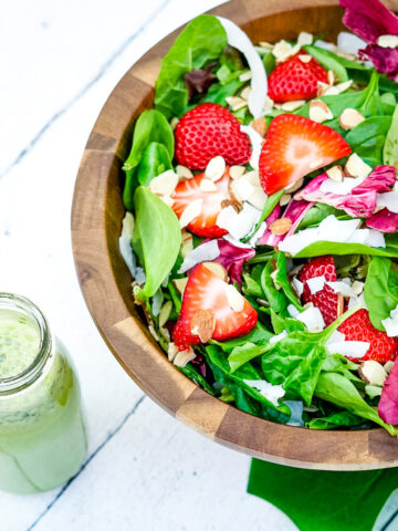 Bright, fresh salad in a wooden bowl with sliced strawberries, red lettuce, shaved coconut and slivered almonds on a white wooden table next to a glass jar of green lemonade basil salad dressing.