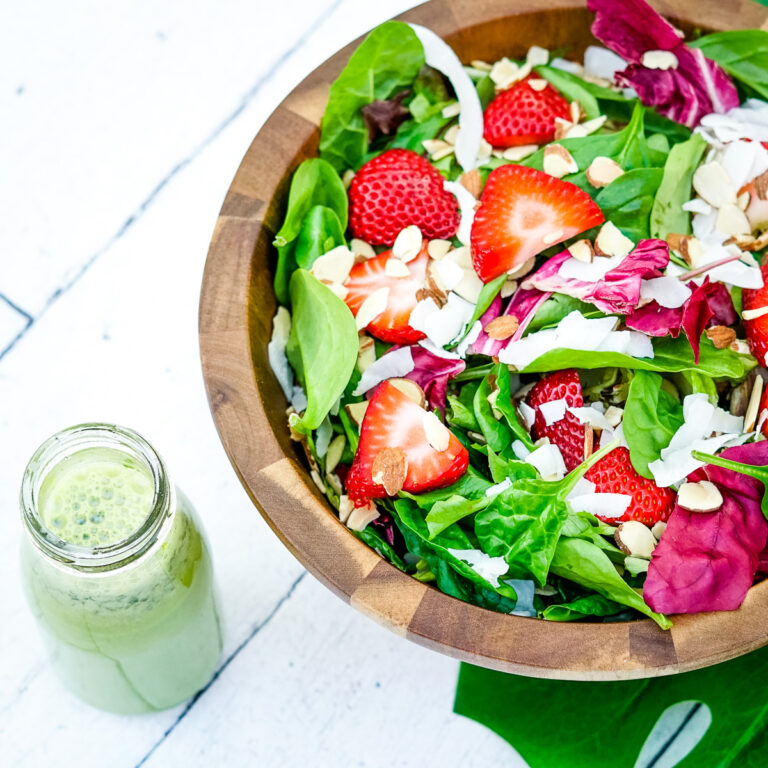 Bright, fresh salad in a wooden bowl with sliced strawberries, red lettuce, shaved coconut and slivered almonds on a white wooden table next to a glass jar of green lemonade basil salad dressing.