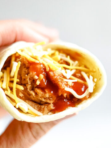 Hand holding a flour tortilla filled with refried beans, taco bell red sauce and shredded cheddar cheese.