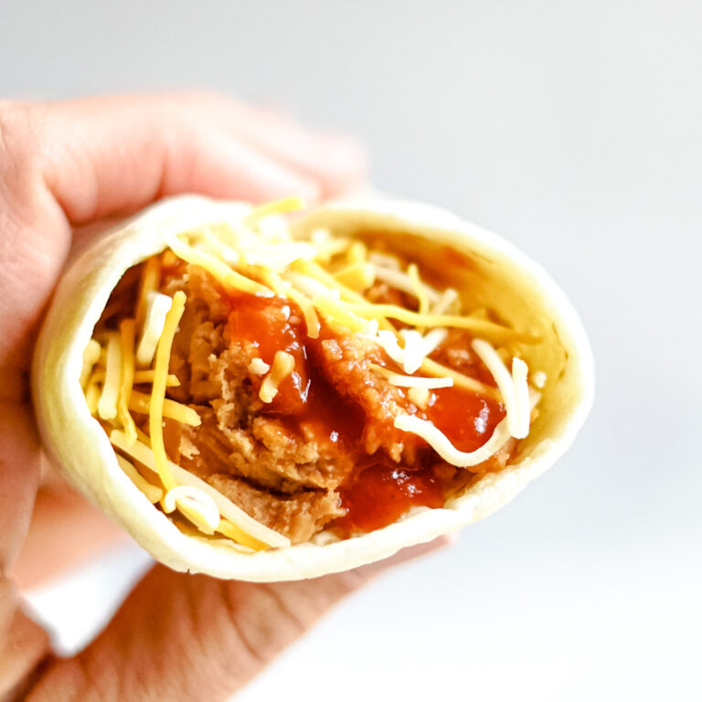 Hand holding a flour tortilla filled with refried beans, taco bell red sauce and shredded cheddar cheese.