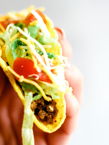Hand holding crispy corn taco shell filled with taco bell seasoned beef, crisp shreds of green lettuce, diced red tomatoes and shredded cheddar cheese.