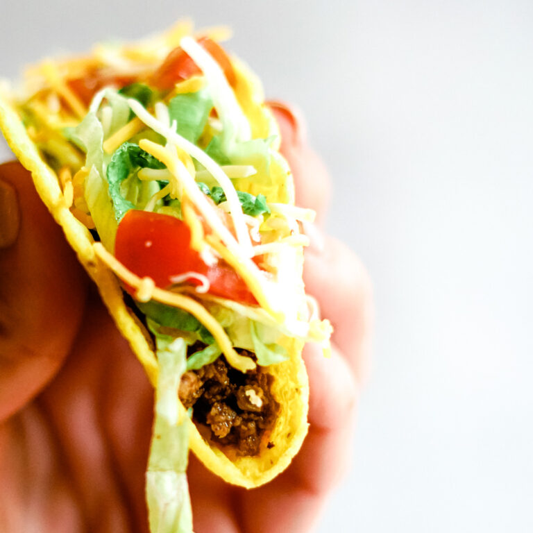 Hand holding crispy corn taco shell filled with taco bell seasoned beef, crisp shreds of green lettuce, diced red tomatoes and shredded cheddar cheese.