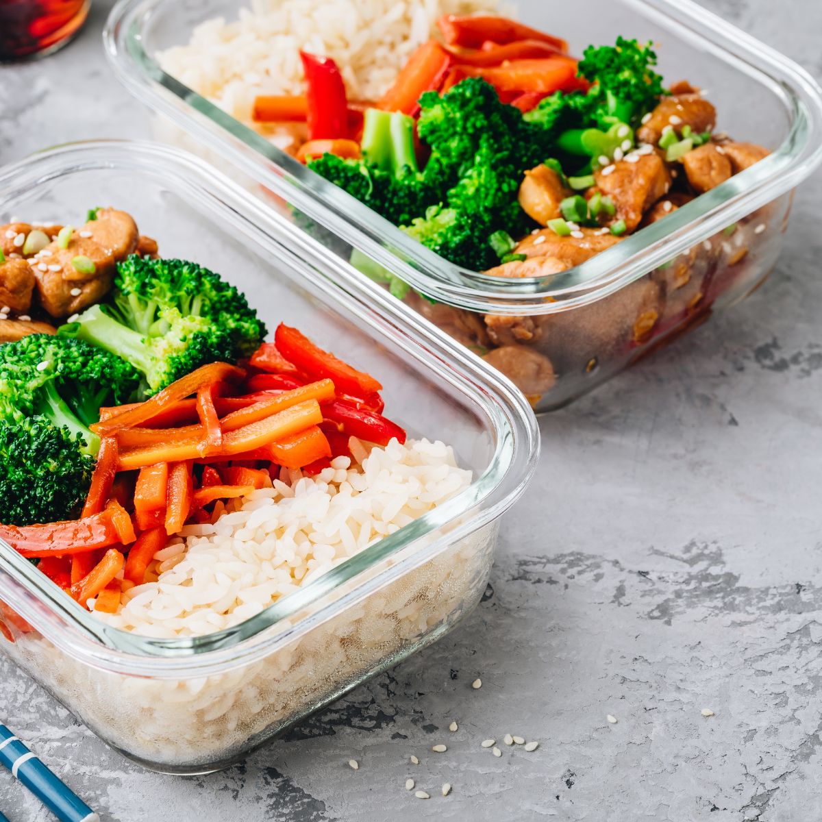 Two hot lunches in glass containers containing white rice, steamed broccoli, sauteed peppers and seasoned chicken with sesame seeds.