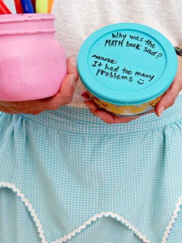 Woman holding markers in one hand and food storage container with silicone lid in the other hand. She's wearing a blue apron. Lid has a joke written on it in sharpie.