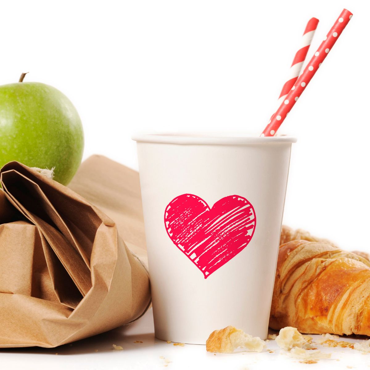 Brown bag lunch with a green apple, croissant and white paper cup with a red heart drawn on it.