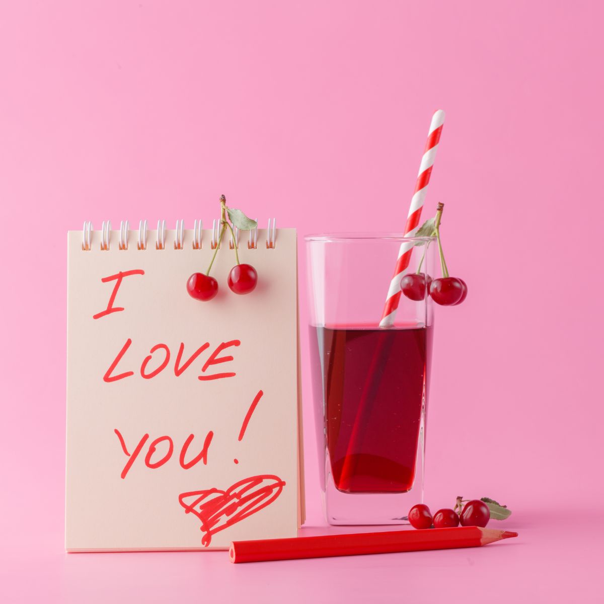 Pink background with red cherry drink with red and white straw. White notepad says I love you in red writing with a heart on it. Cherries strewn about.