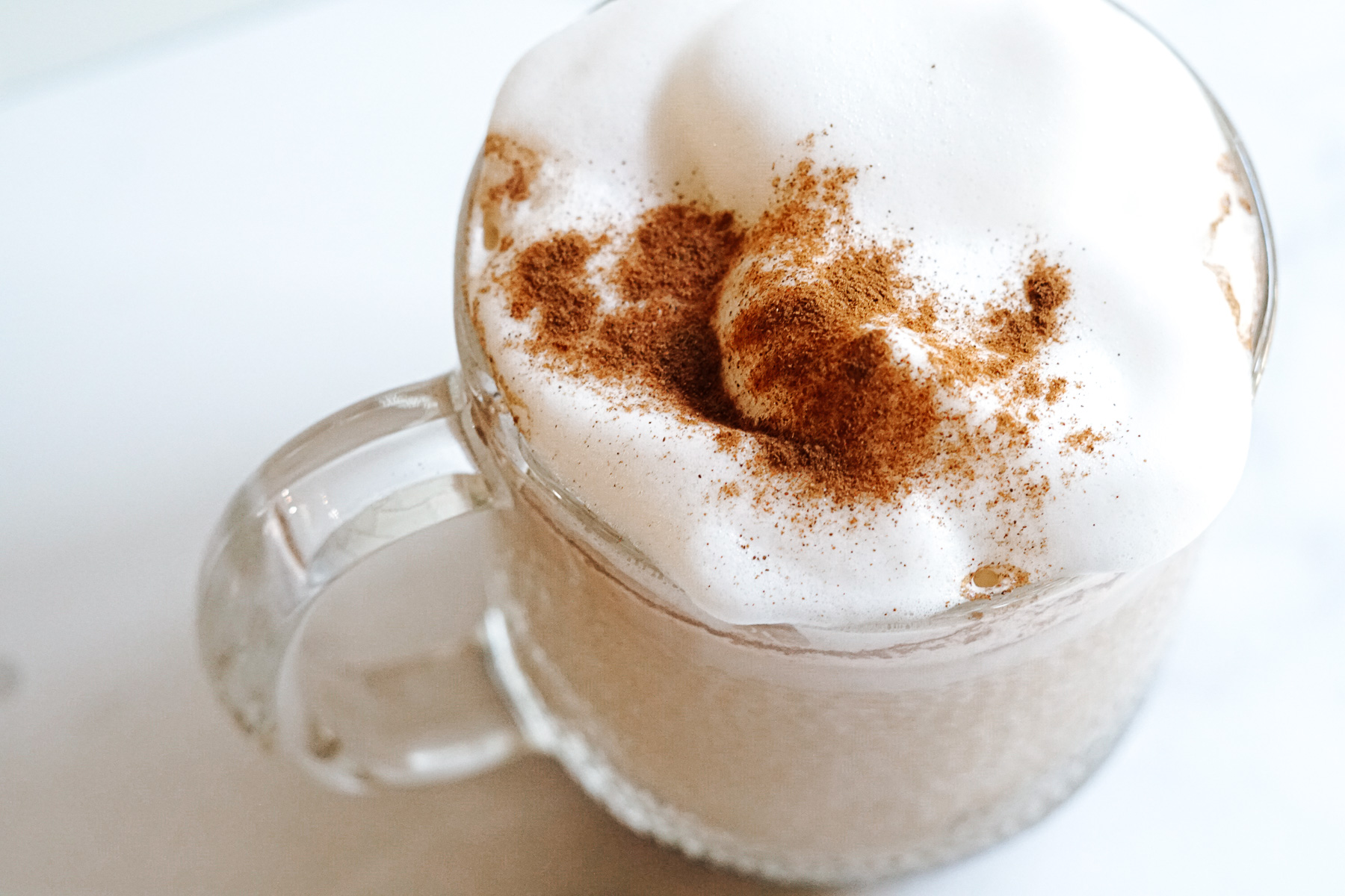 Creamy tan colored coffee drink in clear mug with cold foam and cinnamon on top. Contains coffee, milk and ashwagandha.