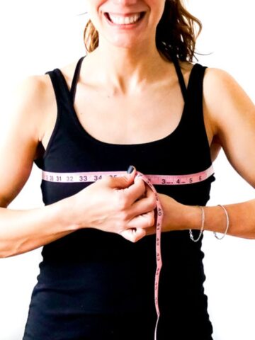 Person wearing a black tanktop and holding a pink fabric tape measure to highlight how to measure your bust. White background.