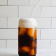 Tall glass filled with ice, cold brew, club soda and cold foam. Glass straw inside.