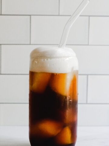 Tall glass filled with ice, cold brew, club soda and cold foam. Glass straw inside.