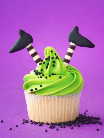 Cupcake with green frosting. Candy that looks like witches legs are sticking out of the top. Purple background.
