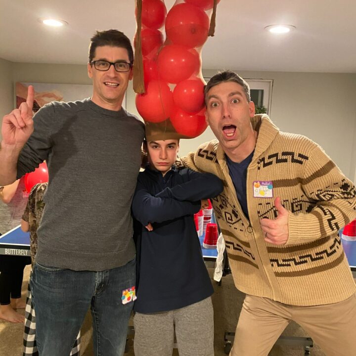 Two dads celebrating winning a game with a child between them wearing panty hose stuffed with balloons on his head.