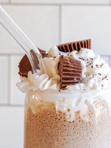 Bailey's peanut butter cup milkshake in a glass topped with whipped cream, full reese's peanut butter cups and a glass straw.