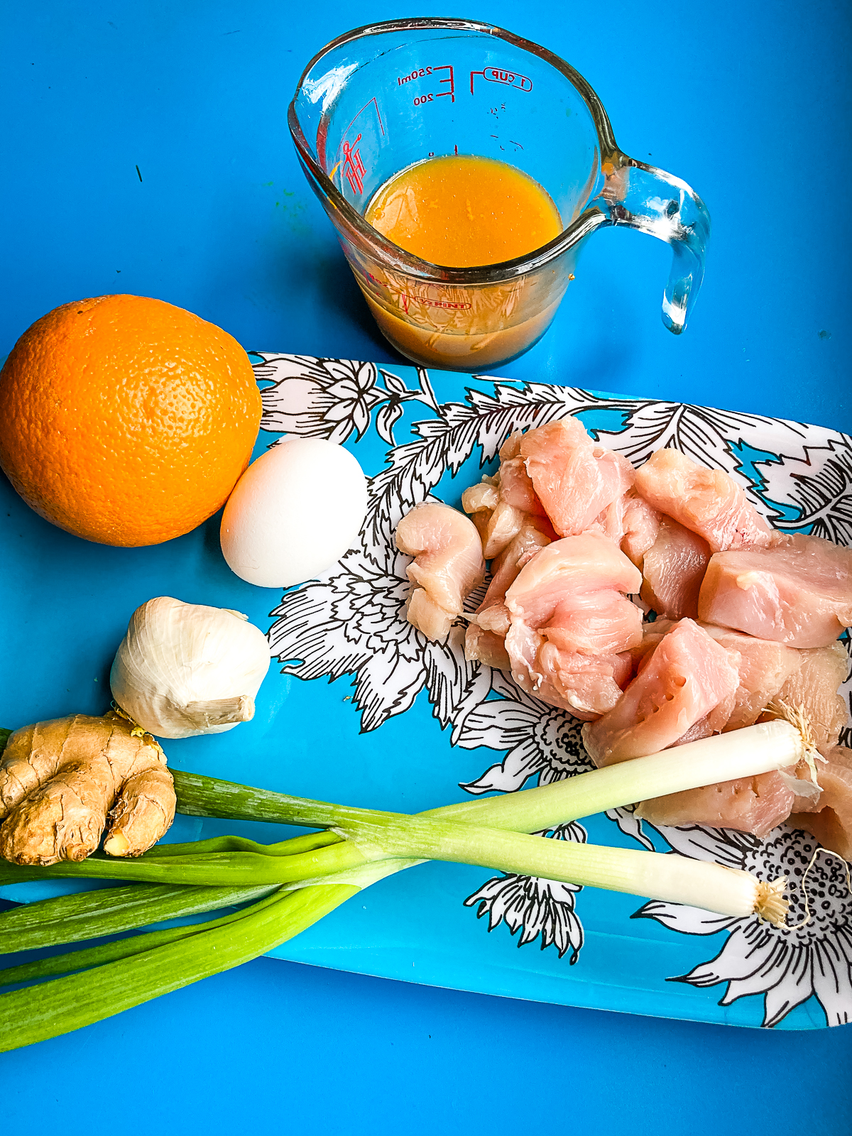ingredients for easy orange chicken on a turquoise plate. Chicken, green onion, ginger, an egg, orange sauce, an orange, and garlic.