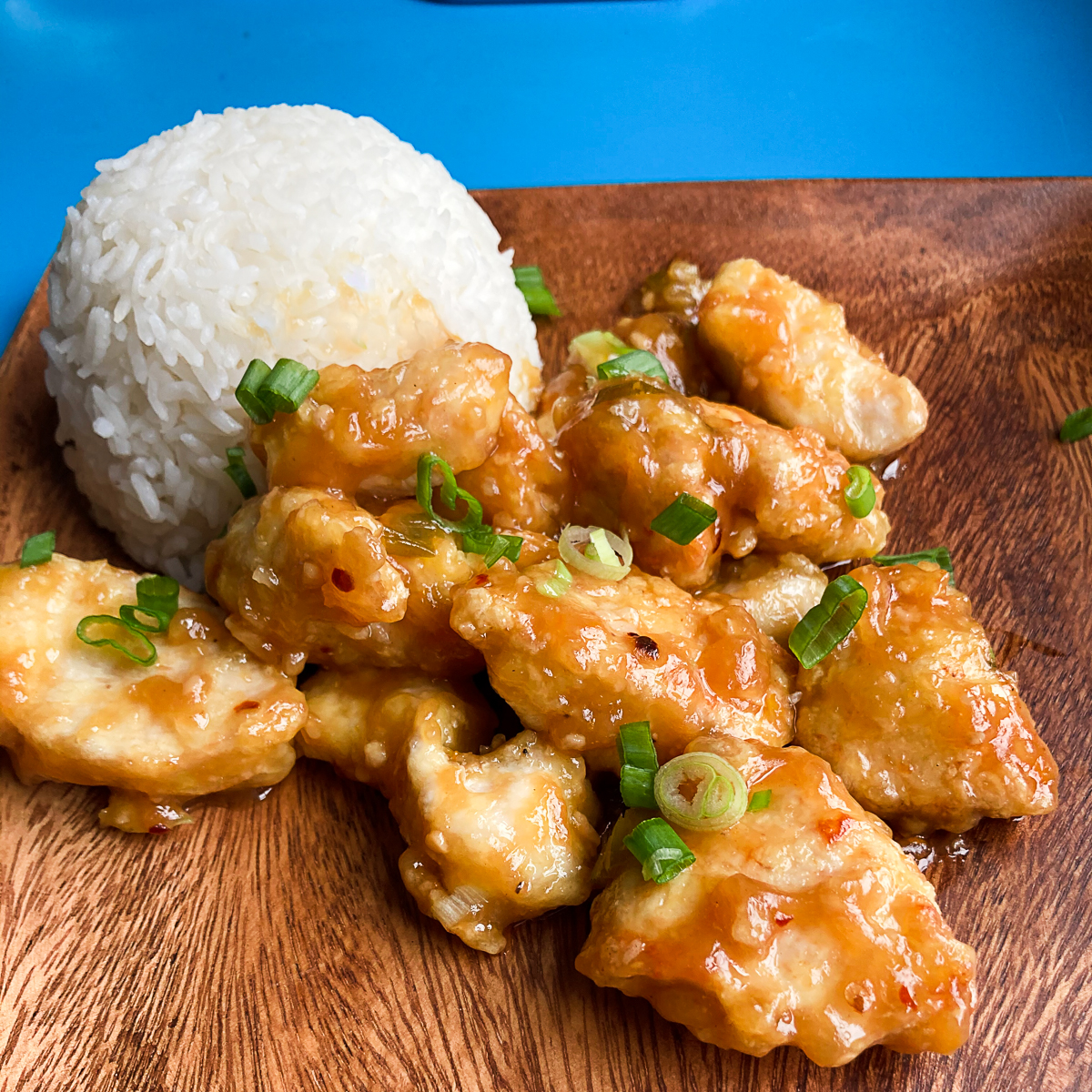 Orange chicken with green onions on a wooden plate. White rice ball on the left side of the plate.