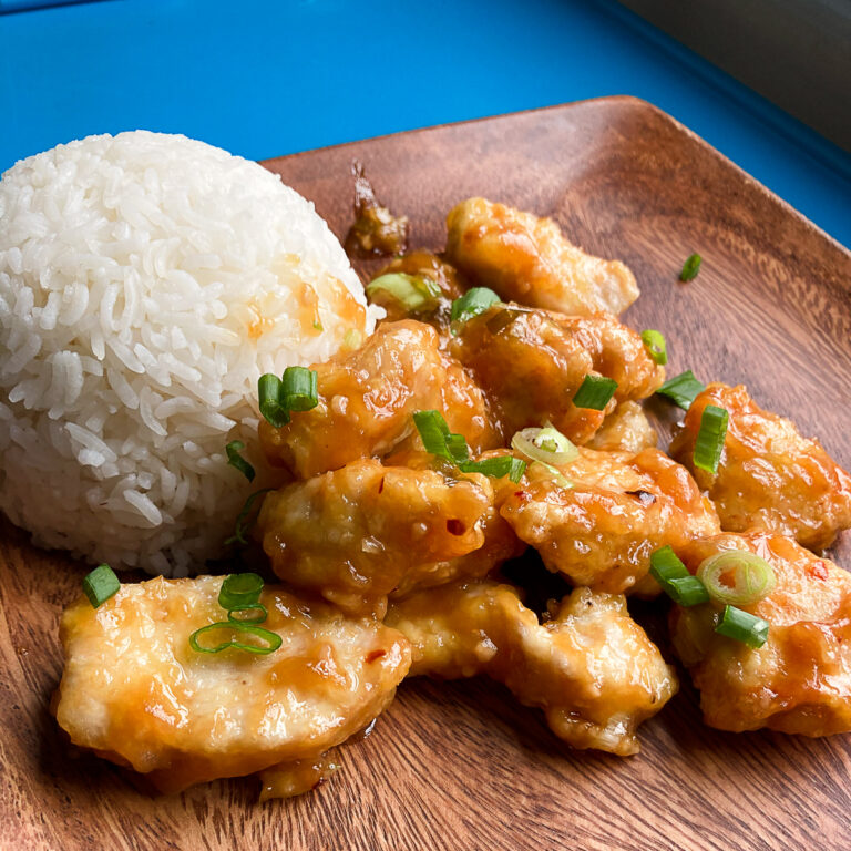 Orange chicken on wooden plate. White rice on side of plate.