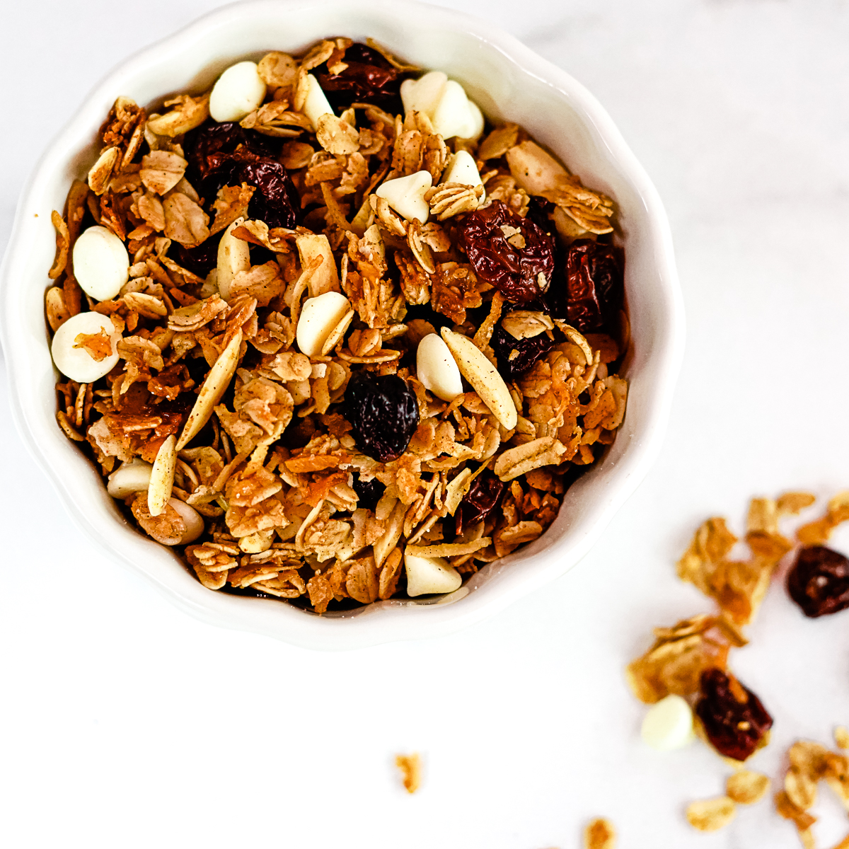 Overhead shot of gingerbread granola in a white bowl on a white countertop. You can see oats, slivered almonds, spices, white chocolate chips and craisins.