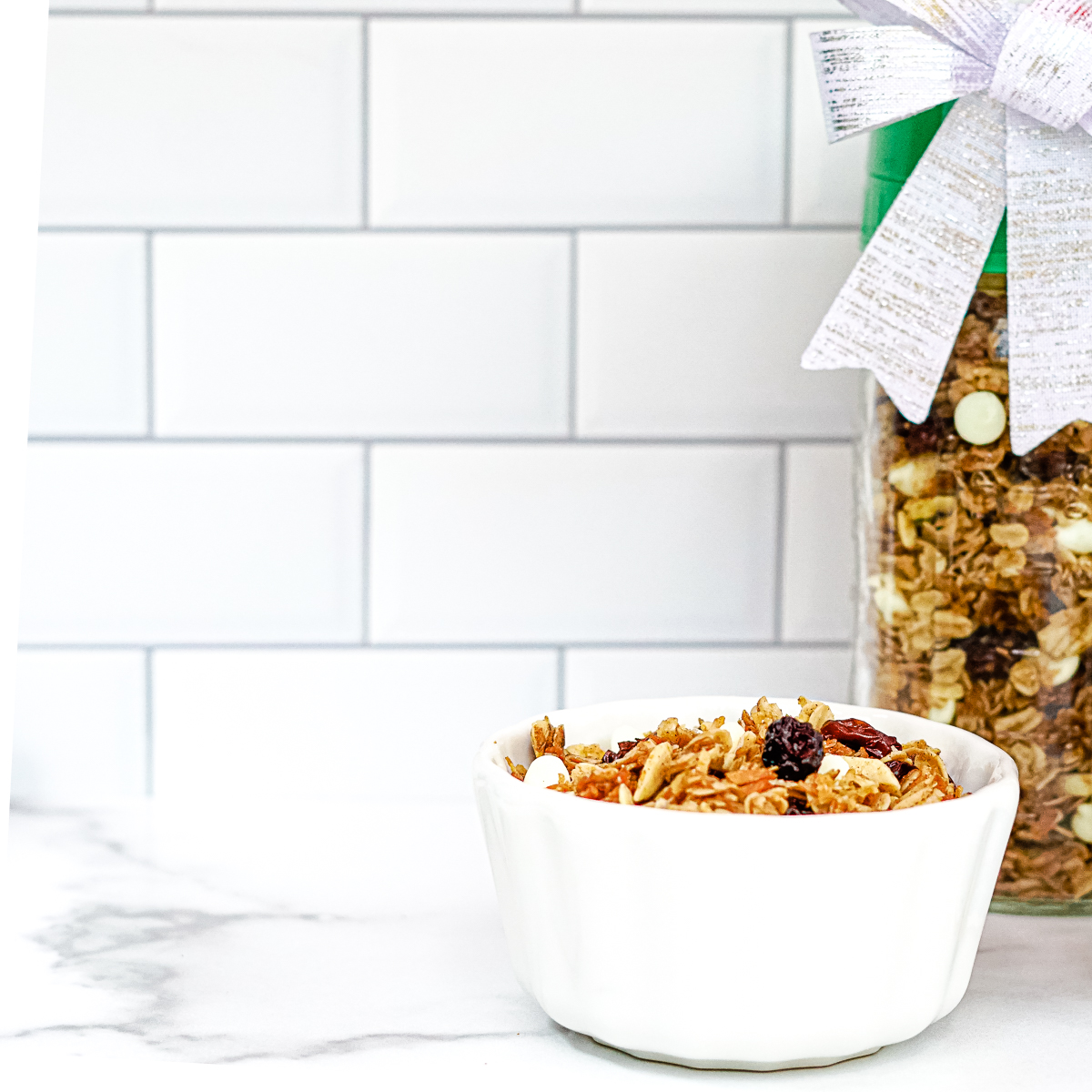 Gingerbread granola in a white bowl on a white countertop. Dried cranberries, oats and white chocolate chips. Jar of granola in the background with a green lid and silver ribbon to be given as a gift.