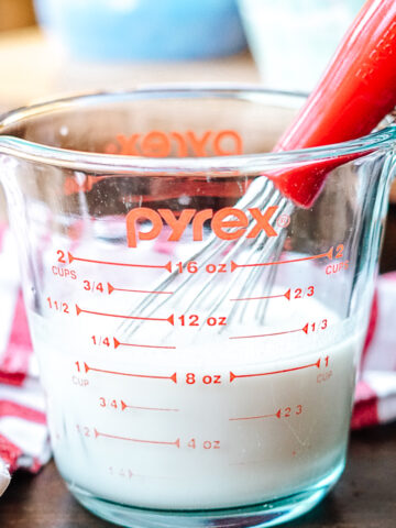Dairy Free Buttermilk in a glass pyrex measuring cup with a red whisk and a red and white striped dish towel in the background
