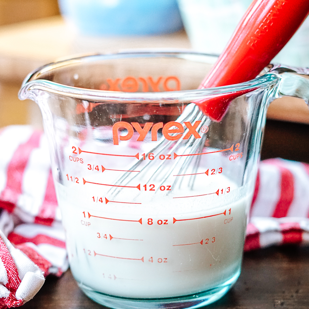 Dairy Free Buttermilk in a glass pyrex measuring cup with a red whisk and a red and white striped dish towel in the background