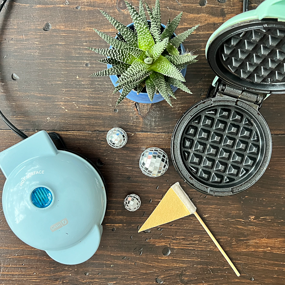 Two Dash Mini Waffle Makers on a wood table. One is closed and one is open. Yellow pennant flag, three small disco balls and a succulent plant are betwen them.