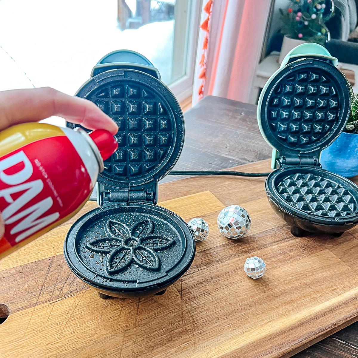 Two Dash Mini Waffle Makers on a wooden cutting board with disco balls between them. Hand holding can of Pam nonstick cooking spray.