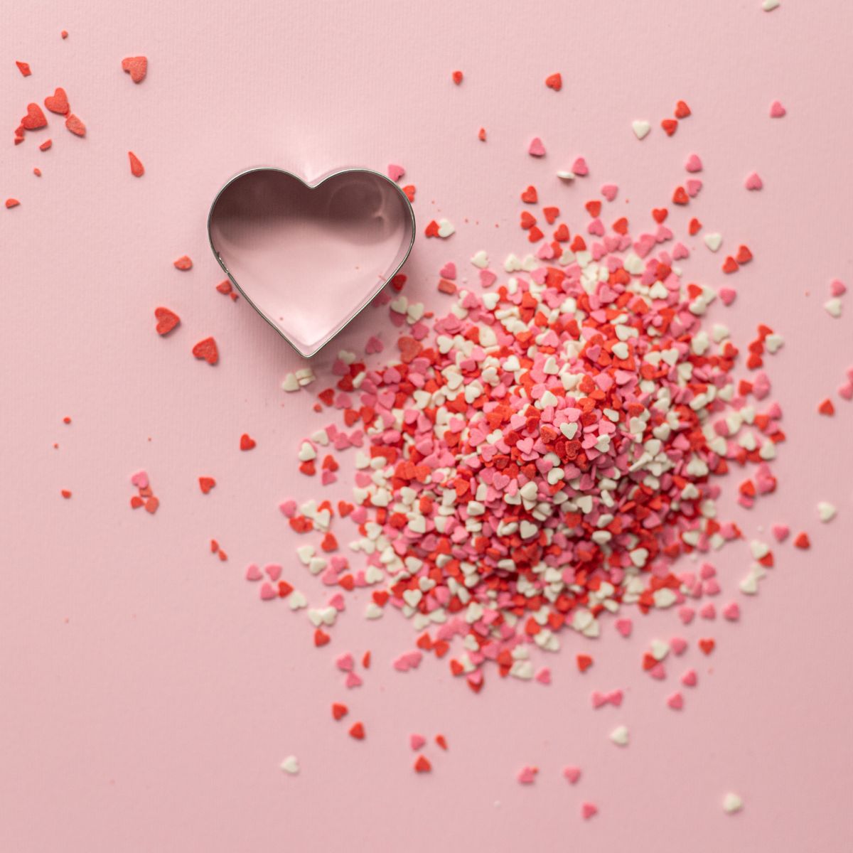 Pink background with small pink, red and white heart shaped sprinkles in a pile and a metal heart shaped cookie cutter next to them.