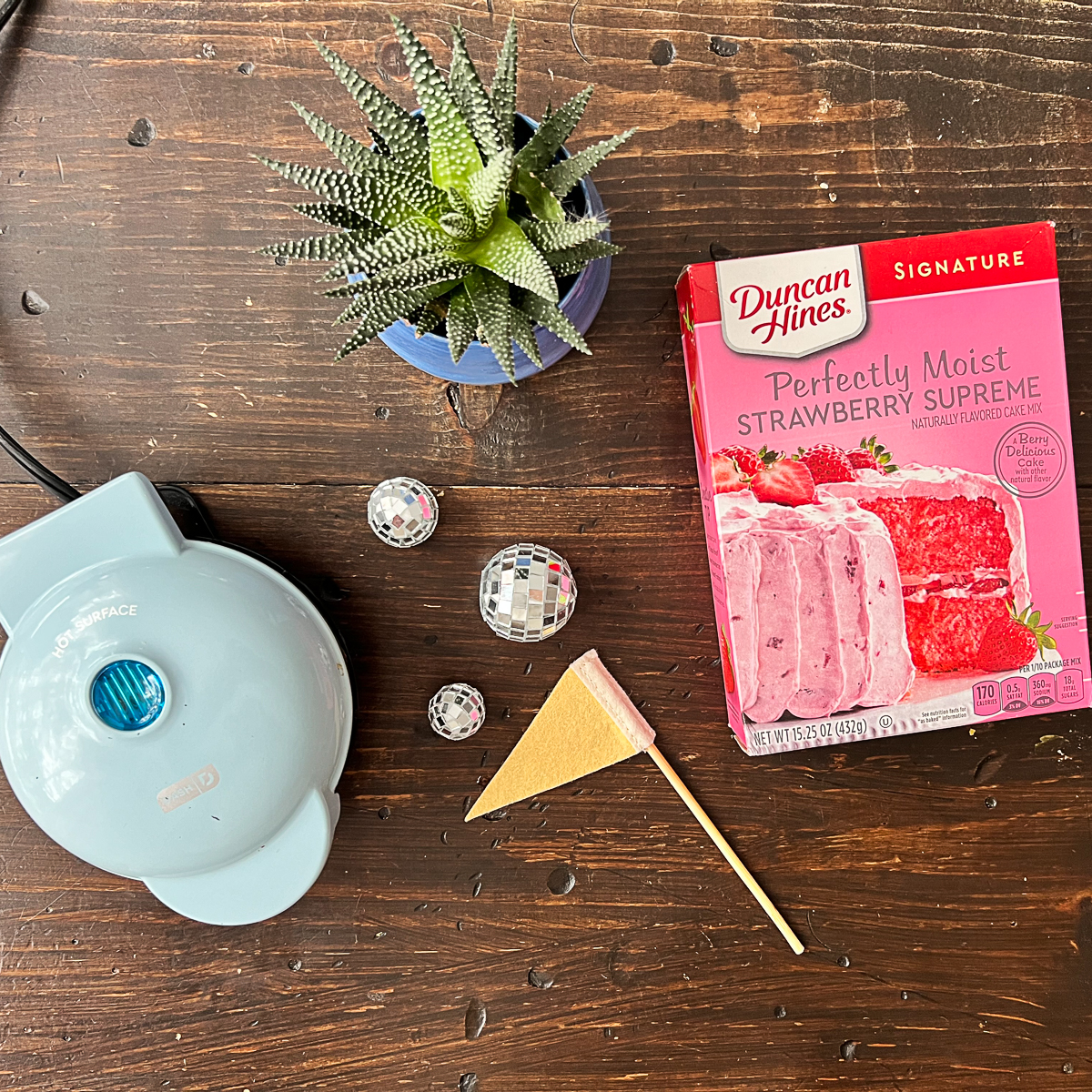 Ingredients for Strawberry cake waffles - box of strawberry cake mix and a Dash Mini Waffle Maker with a pennant flag, succulent and three small disco balls on a dark table